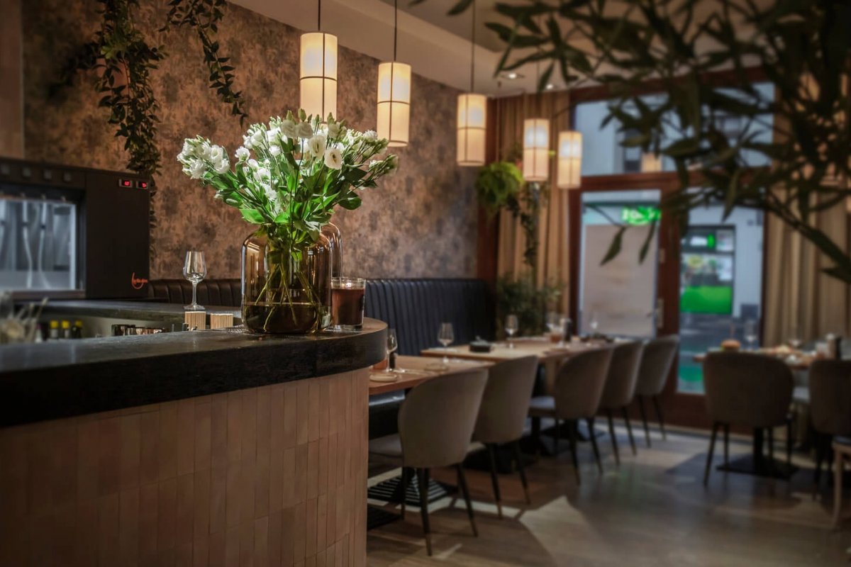 Wnętrze eleganckiej restauracji z nastrojowym oświetleniem, kwiatami w szklanym wazonie na barze i starannie nakrytymi stolikami w stylu nowoczesnym.